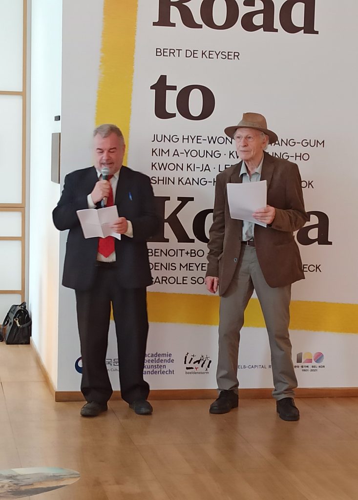 The two protagonists standing before a poster for the exhibition