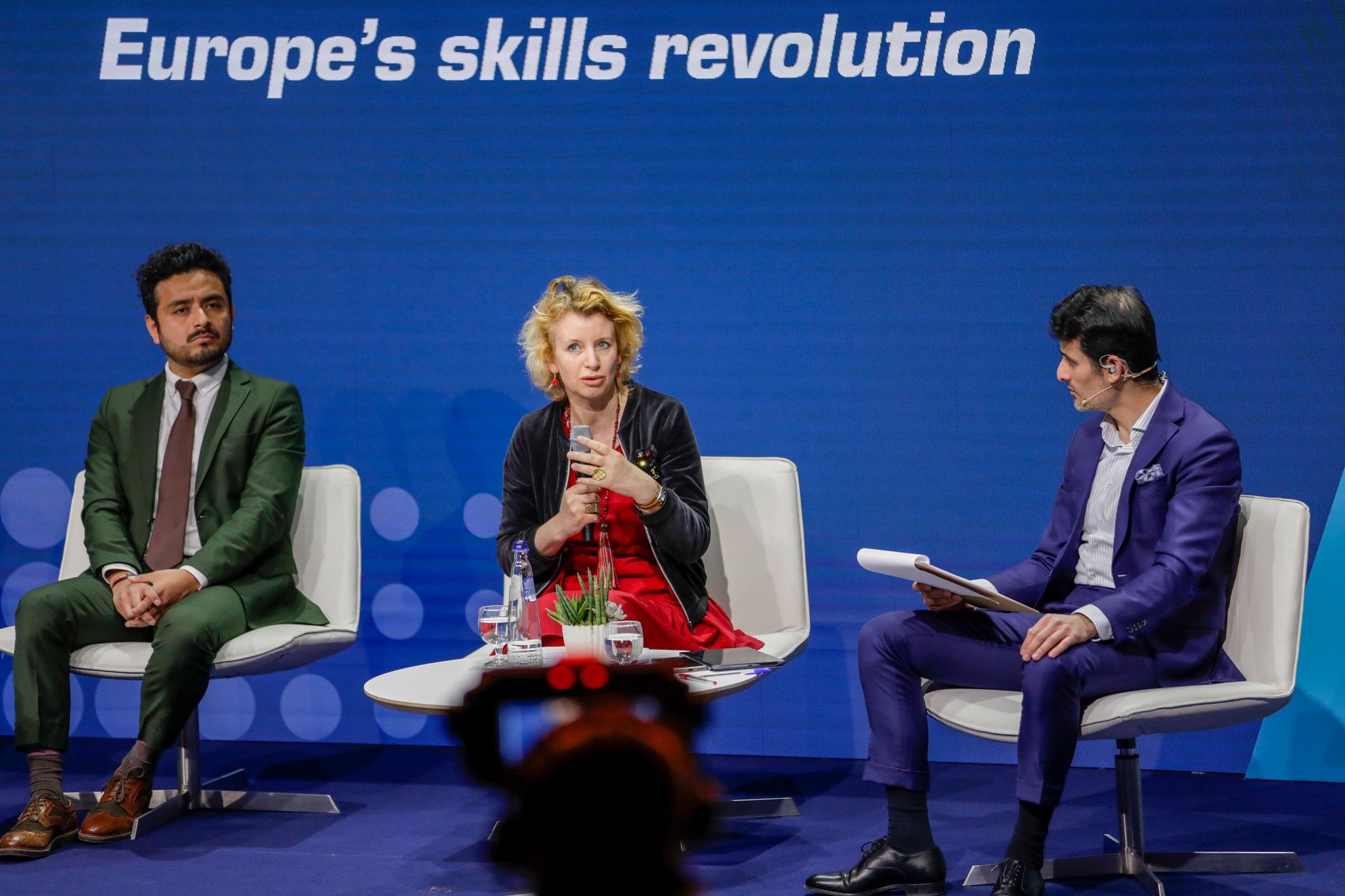 Trois personnes se trouvent sur un podium, l’une d’eux, une dame blonde, tient un micro