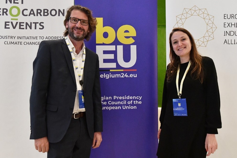 A man and a woman are posing in front of a banner of the Belgian presidency