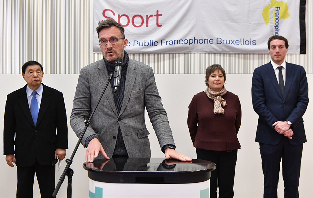 The State Secretary, standing behind a lectern with a microphone, flanked by three people, in front of a banner of the French Community Commission. 