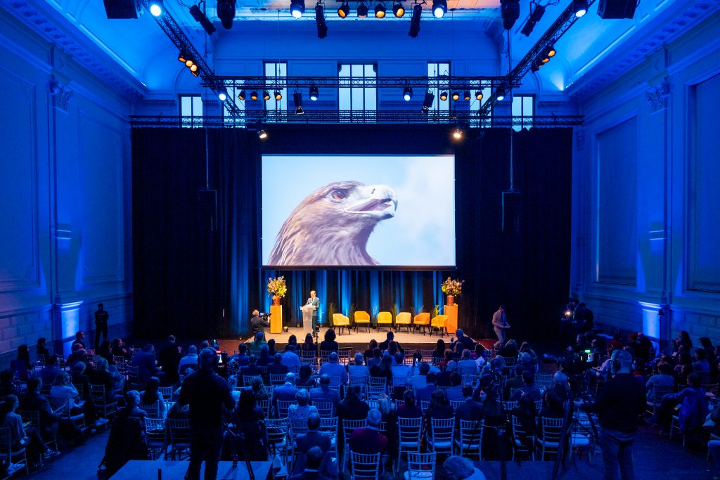 Een zaal vol zittende toeschouwers. Op het podium staat een een scherm waar het hoofd van een roofvogel te zien is.