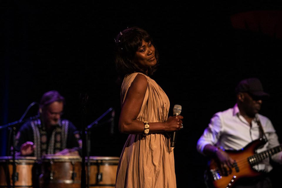 the singer standing, holding a microphone in one hand, flanked by a percussionist and a bass player