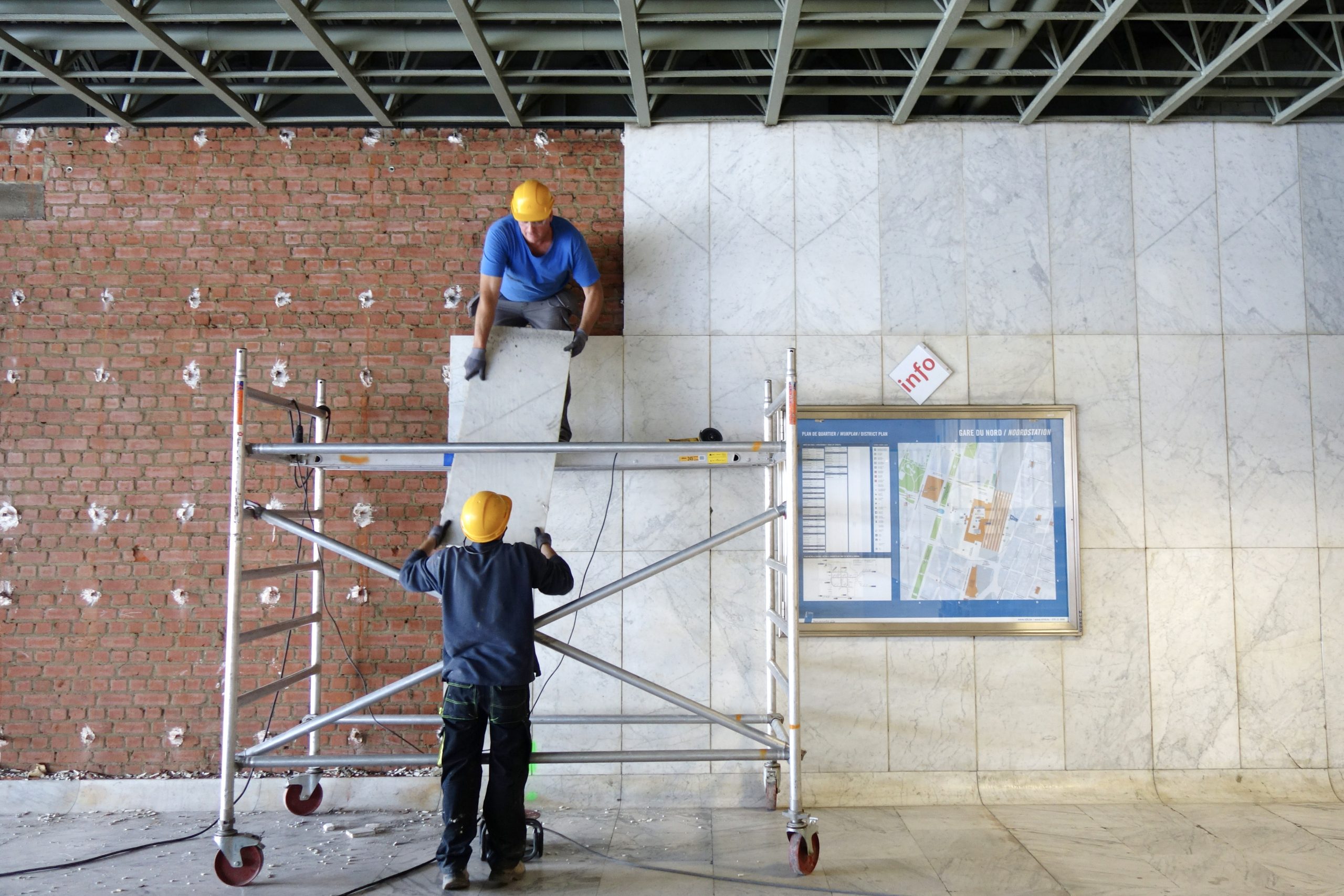 Two workers, on a scaffold, are working on covering a wall.