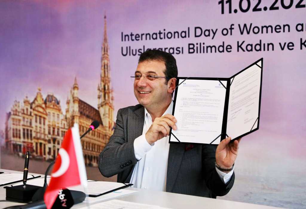 The Mayor, seated at a desk, holds up the signed document.