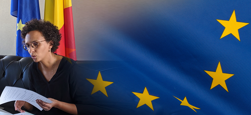La Secrétaire d’État parle avec une feuille en main, assise devant les drapeaux européen et belge
