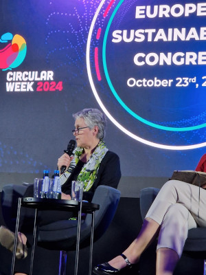 Une dame, assise sur scène, tient un micro et participe à un panel au European Sustainability Congress le 23 octobre 2024. Le logo de l'événement « Circular Week 2024 » est visible en arrière-plan.