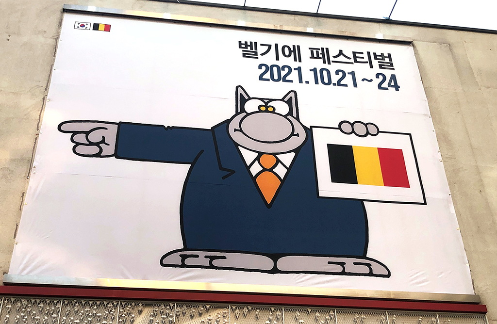 Exhibition poster featuring Le Cat pointing in one direction, holding the Belgian flag in his other hand.