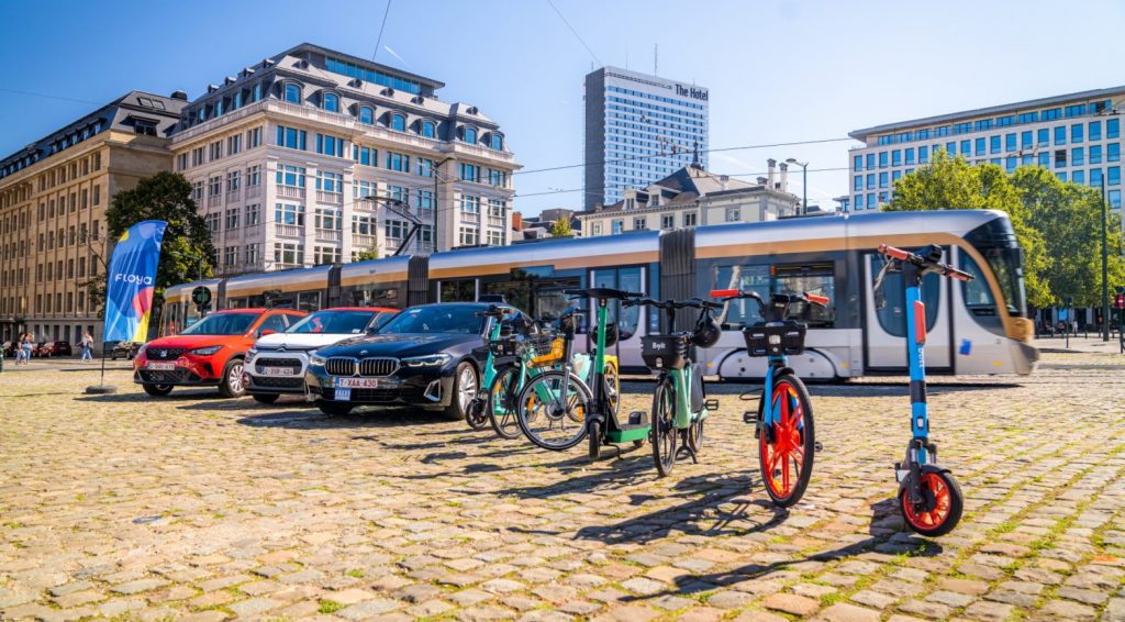 A beach flag, three cars, bicycles and scooters in front of a STIB-MIVB tram.